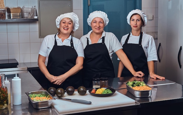 Gratis foto gelukkig kookteam in uniform naast een tafel met gehakte groenten, poserend voor een camera in de keuken van een restaurant.