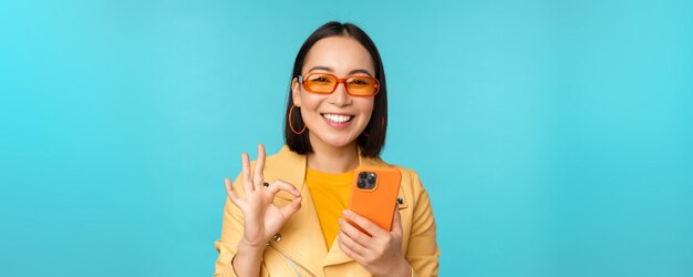Gelukkig koreaans meisje met een zonnebril die een goed teken toont en een mobiele telefoon vasthoudt met behulp van een smartphone-app die een toepassing aanbeveelt die over een blauwe achtergrond staat