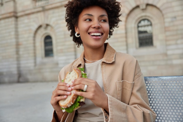 Gelukkig krullend haired vrouw zit buiten eet heerlijke sandwich kijkt graag weg geeft de voorkeur aan junkfood gekleed in casual kleding heeft lunchpauze na een wandeling in de stad mensen levensstijl voeding