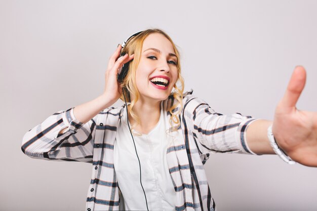 Gelukkig krullend meisje in gestreept overhemd glimlachen en dansen tijdens het luisteren naar favoriete liedje in oortelefoons. Close-up portret van charmante jonge vrouw in koptelefoon met plezier