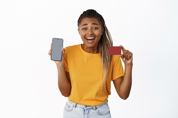 Gelukkig lachend Afrikaans-Amerikaans meisje toont creditcard en smartphonescherm met een gele, casual t-shirt op een witte achtergrond