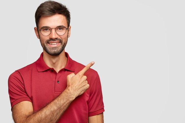Gelukkig lachend Europese man met borstelharen, wijst met wijsvinger opzij, nodigt u uit om te dineren in een restaurant in de buurt, gekleed in een casual rood t-shirt, bril geïsoleerd op een witte muur