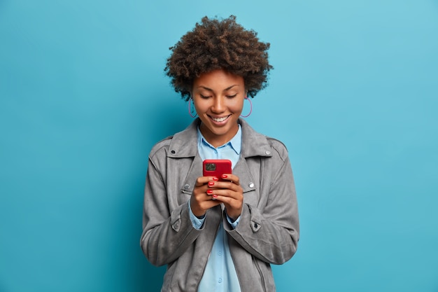 Gelukkig lachend gekrulde haired jonge vrouw typen bericht op mobiele telefoon, kijkt met blije uitdrukking op het display, draagt grijze jas,