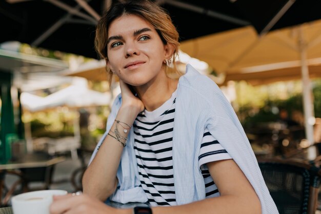 Gelukkig lachende Europese vrouw die een gestreept t-shirt draagt, rust op een open luchtterras in het zonlicht op een warme lentedag Ochtendkoffie op een zonnige dag