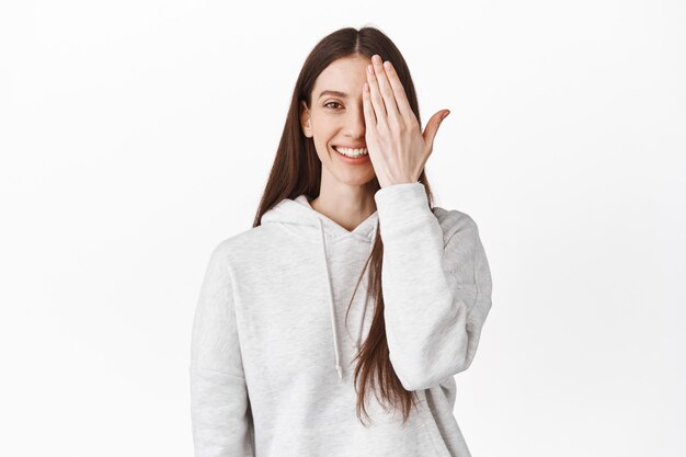 Gelukkig lachende jonge vrouw bedek de helft van het gezicht, kijkend naar de voorkant met één kant, staande in een casual hoodie tegen een witte muur