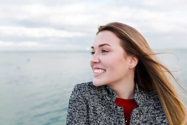 Gelukkig lachende langharige vrouw met grote blauwe ogen in rood shirt en grijze jas loopt in de buurt van de zee