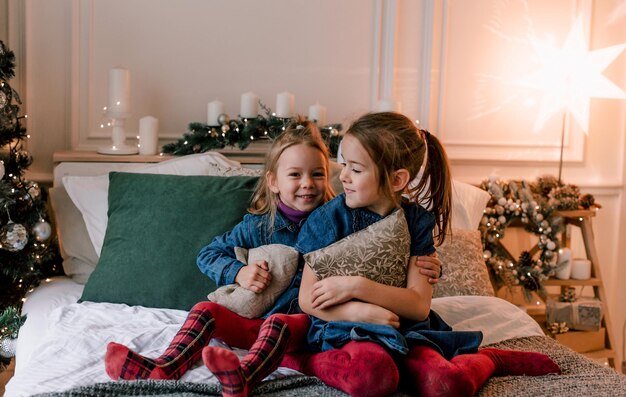 Gelukkig lachende meisjes zitten op het bed en knuffelen kussens in hun handen op kerstavond