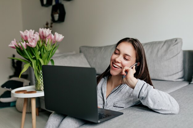 Gelukkig lachende vrouw in pyjama praten aan de telefoon en werken met laptop in modern, stijlvol appartement in quarantaine.