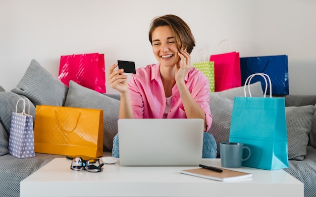 Gelukkig lachende vrouw in roze shirt op de bank thuis onder kleurrijke boodschappentassen met creditcard online betalen op laptop