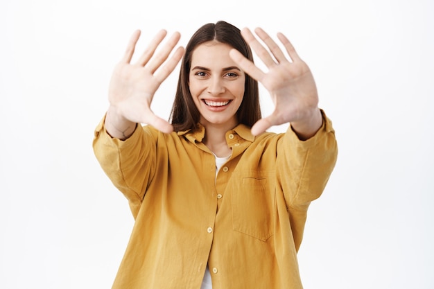 Gelukkig lachende vrouw strekt de handen naar voren en kijkt door dromerig, iets moois voorstellend, een interessant concept creërend, staande over een witte muur