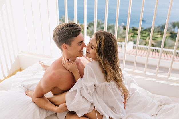 Gelukkig langharige meisje omhelst zachtjes haar lachende vriendje en gaat hem kussen. Mooie jonge paar schattig knuffelen en lachen, terwijl rusten in bed in zonnige ochtend op zee resort