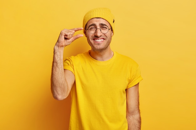 Gelukkig man in gele casual kleding, maakt een klein gebaar, toont iets heel kleins, lacht graag, heeft een vrolijke uitdrukking, draagt een transparante bril