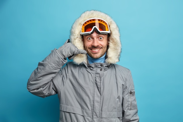 Gelukkig mannelijke snowboarder draagt winterjas met capuchon maakt telefoongebaar positief glimlacht.