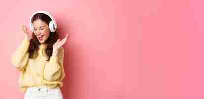 Gratis foto gelukkig meisje dat haar favoriete liedje zingt en naar muziek luistert in een draadloze koptelefoon die lacht en danst over roze achtergrond