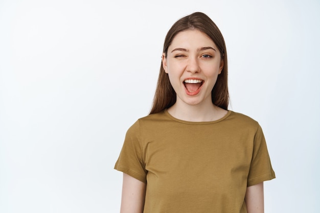 Gelukkig meisje glimlachend, knipogend naar de camera. Vrouw heeft een witte perfecte glimlach en een schone huid, staat in een casual t-shirt op een geïsoleerde studio.