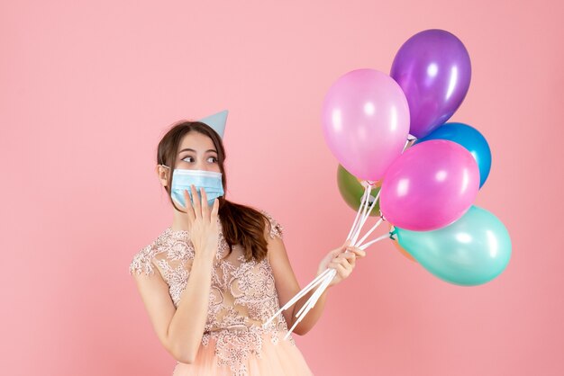 gelukkig meisje met feestmuts en medisch masker met kleurrijke ballonnen op roze