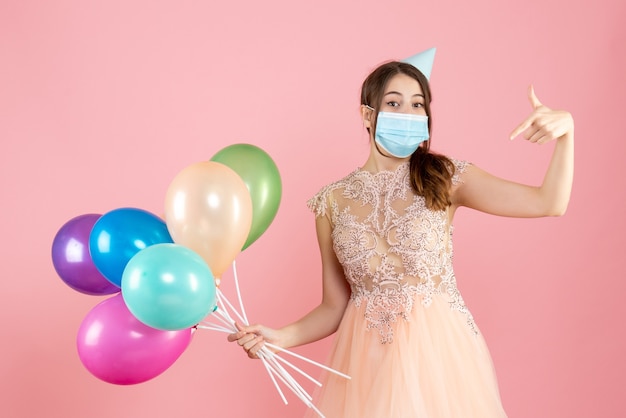 gelukkig meisje met feestmuts en medisch masker wijzend naar zichzelf met kleurrijke ballonnen op roze