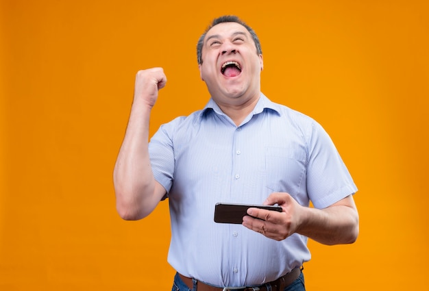 Gelukkig middelbare leeftijd man met blauwe gestripte shirt winnende spel op mobiele telefoon en zijn hand opheft in het gebaar van triomf terwijl hij staat