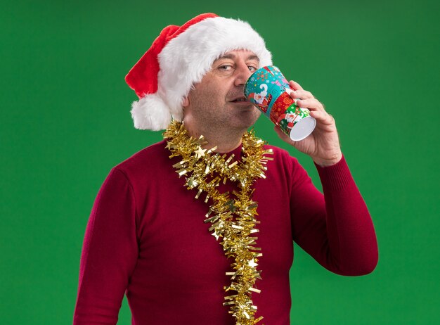Gelukkig middelbare leeftijd man met kerst kerstmuts met klatergoud rond de nek drinken uit kleurrijke papieren beker staande over groene achtergrond