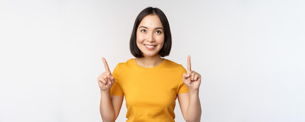 Gelukkig mooi Aziatisch meisje dat reclame toont die met vingers omhoog wijst die over witte achtergrond staat