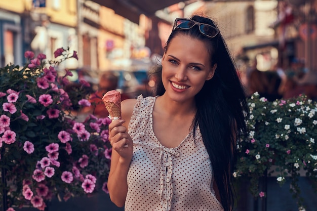 Gelukkig mooi brunette meisje dat trendy kleding draagt, geniet van een zomerdag met aardbeienijs terwijl ze in de buurt van een met bloemen versierd terras staat.