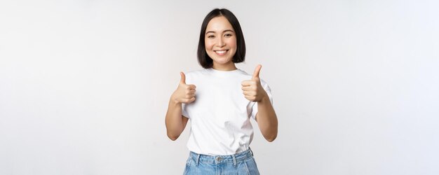 Gelukkig mooi Koreaans meisje dat duimen omhoog laat zien in goedkeuring glimlachend tevreden als iets dat aanbeveelt om op een witte achtergrond te staan