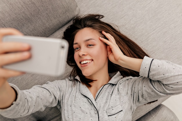 Gelukkig mooi meisje met behulp van smartphone in de ochtend en selfie maken. indoor portret van prachtige donkerharige vrouw ontspannen thuis.