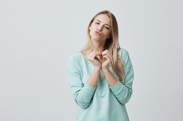 Gelukkig mooi wijfje met blond lang haar die liefdetekens met haar handen tonen die in hartvorm tot een kom worden gevormd. Blanke vrouw verliefd pruilende lippen, kusjes verzenden, positieve emoties uitstralen.