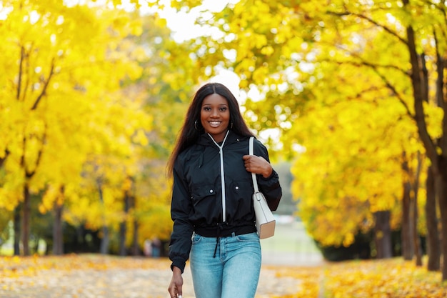 Gelukkig mooie afrikaanse lachende vrouw met zwarte huid in mode kleding met jas, jeans en handtas wandelingen in het herfstpark met gele herfstbladeren