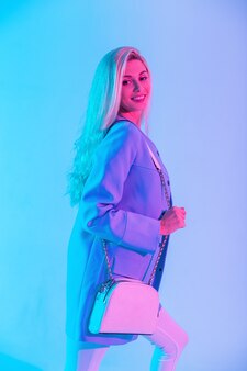 Gelukkig mooie glimlachende zakenvrouw met mooi vrouwelijk gezicht in modieuze blauwe jas met witte leren handtas op neon roze licht in studio