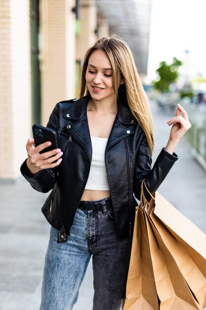 Gelukkig mooie jonge vrouw met boodschappentassen, maakt gebruik van een mobiele telefoon, staande in de stad