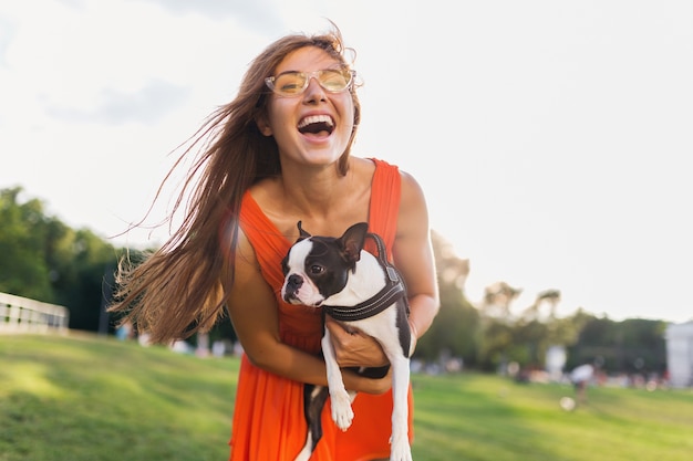 Gelukkig mooie vrouw park bedrijf Boston Terriër hond, glimlachend positieve stemming, trendy zomerstijl, oranje jurk, zonnebril dragen, spelen met huisdier, plezier maken, zonnig weekendentertainment