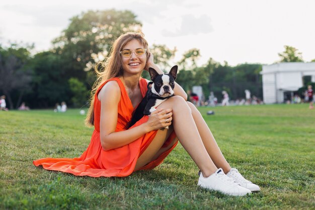 Gelukkig mooie vrouw zittend op het gras in zomer park, boston Terriër hond houden, glimlachend positieve stemming, oranje jurk, trendy stijl, slanke benen, sneakers, spelen met huisdier, ontspannen