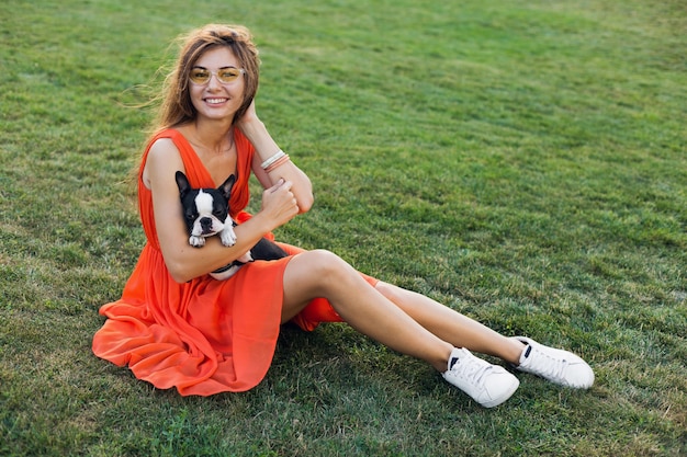 Gelukkig mooie vrouw zittend op het gras in zomer park, boston Terriër hond te houden, glimlachend positieve stemming, oranje jurk, trendy stijl, slanke benen, sneakers, spelen met huisdier