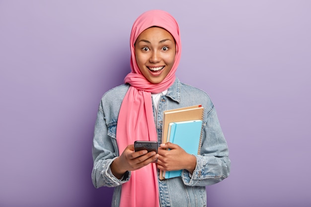 Gelukkig moslimvrouw geavanceerde gebruiker van technologie houdt blocnotes en mobiele telefoon, draagt een roze sluier op het hoofd