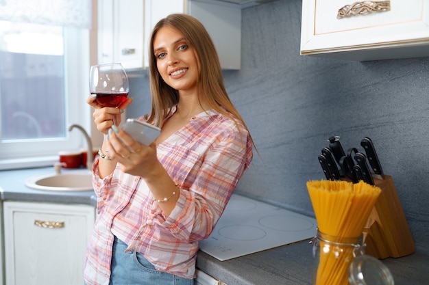 Gelukkig ontspannen jonge vrouw permanent in de keuken met glas rode wijn en met behulp van haar smartphone voor videocall