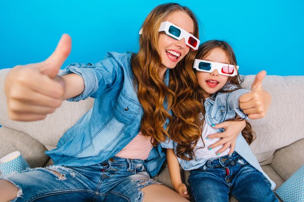 Gelukkig opgewonden moeder knuffelen schattige mooie dochter op Bank op blauwe achtergrond. Samen 3D-films kijken in een bril, jeanskleding dragen, positiviteit en geluk uitdrukken voor de camera