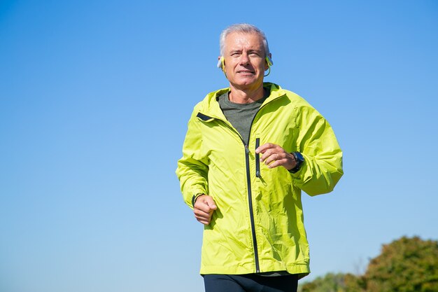 Gelukkig opgewonden senior man in draadloze koptelefoon joggen buiten. Lage hoek, blauwe heldere lucht. Vooraanzicht, kopieer ruimte. Activiteit en leeftijd concept