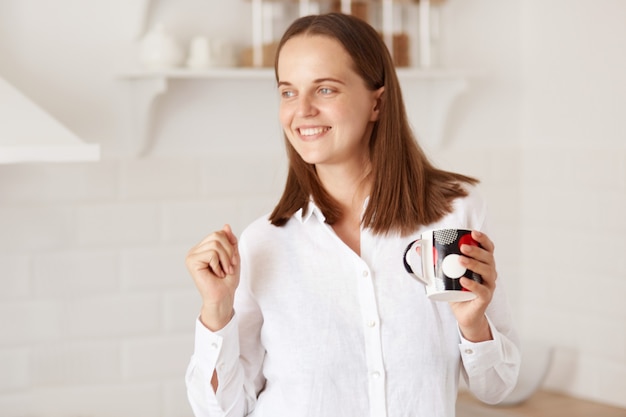 Gelukkig opgewonden vrouw die vroeg in de ochtend wakker wordt, met een kopje koffie of thee in handen staat en danst, een goed humeur heeft, poserend met een keuken op de achtergrond.