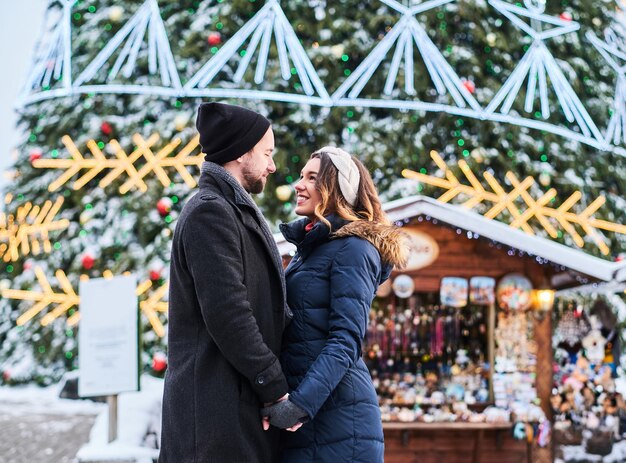 Gelukkig paar dat warme kleren draagt, houdt elkaars hand vast en kijkt elkaar aan, staat in de buurt van een stadskerstboom en geniet ervan om samen tijd door te brengen.