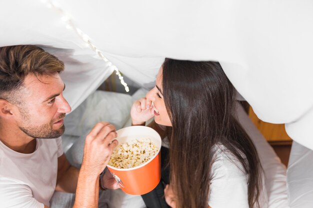 Gratis foto gelukkig paar die elkaar bekijken die popcorn zittend op bed eten