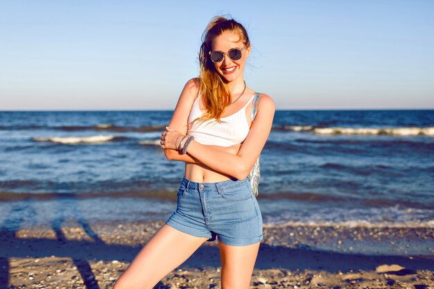 Gelukkig positief portret van jonge blonde vrouw plezier op tropische vakantie, wandelen in de buurt van de oceaan, reizen met rugzak, stijlvolle hipster zomer outfit en zonnebril. gebruind sexy lichaam, lange haren.