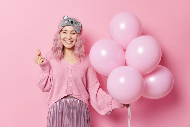 Gelukkig positieve Aziatische vrouw maakt Koreaans als teken vormen mini hart drukt liefde houdt bos ballonnen draagt slaapmasker trui en rok viert verjaardag geïsoleerd over roze achtergrond