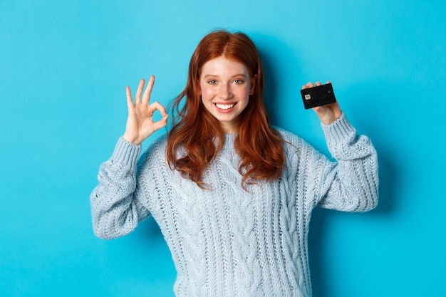 Gelukkig roodharig meisje in trui met creditcard en goed teken, bankaanbieding aanbevelend, staande over blauwe achtergrond