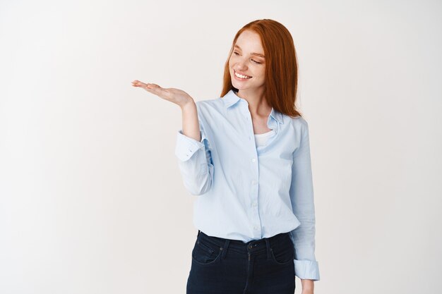 Gelukkig roodharige vrouw met speciale promotie bij de hand, kijkend naar product met een tevreden glimlach, staande over een witte muur