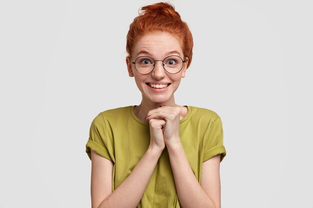 Gelukkig roodharige vrouw met sproeten en positieve glimlach houdt de handen bij elkaar, verwacht iets verrassends, draagt een casual t-shirt