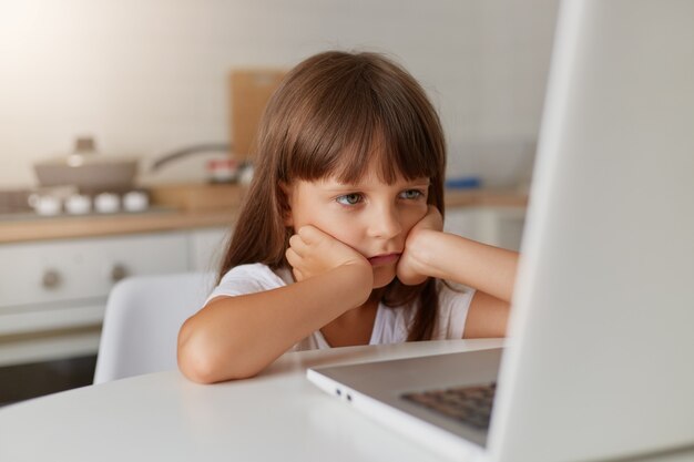 Gelukkig schattig klein meisje van 8 jaar oud in een gestreept t-shirt en jeans met bril zit thuis op een tapijt voor een laptop, onderwijstechnologieën op afstand en huiswerk