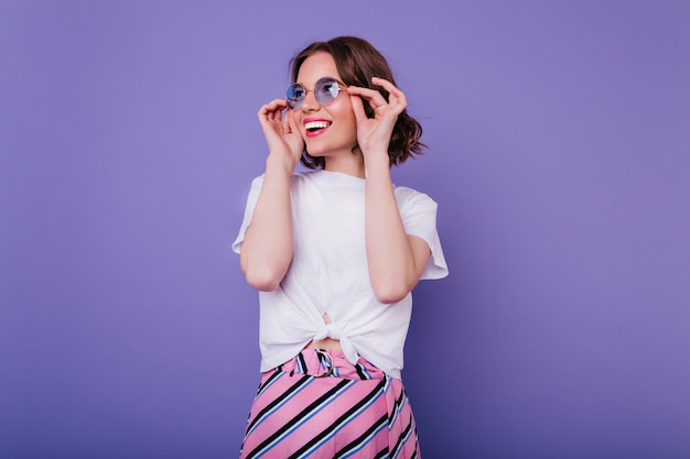 Gelukkig schattig meisje met golvend kapsel haar bril met glimlach aan te raken. Binnen schot van prachtige krullende dame in wit t-shirt poseren op paarse muur.