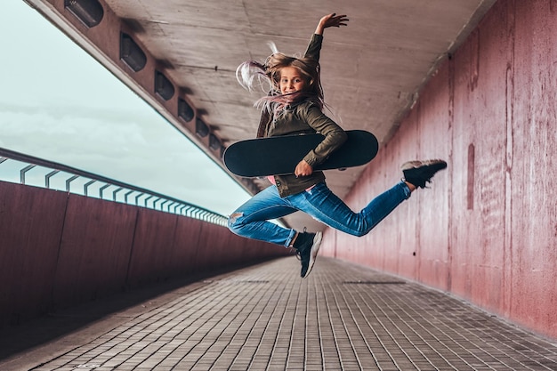 Gratis foto gelukkig schoolmeisje met blond haar gekleed in trendy kleding springt met een skateboard op het voetpad van de brug.