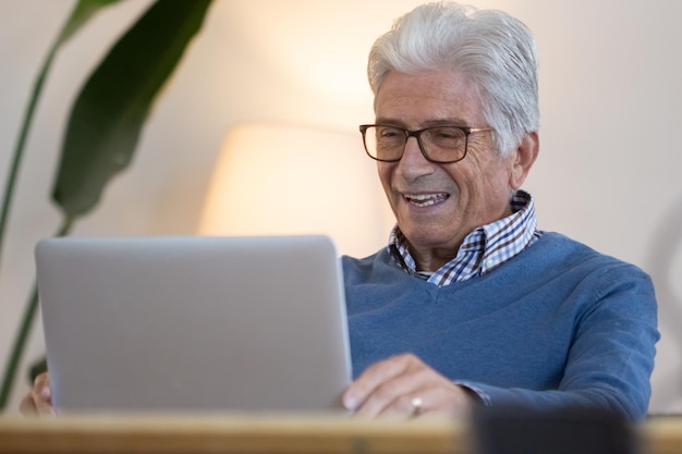 Gelukkig senior man met videogesprek met vrienden thuis. Lachende volwassen man in bril zittend aan tafel in de woonkamer, scherm kijken en praten. Groet, concept van digitale apparaten